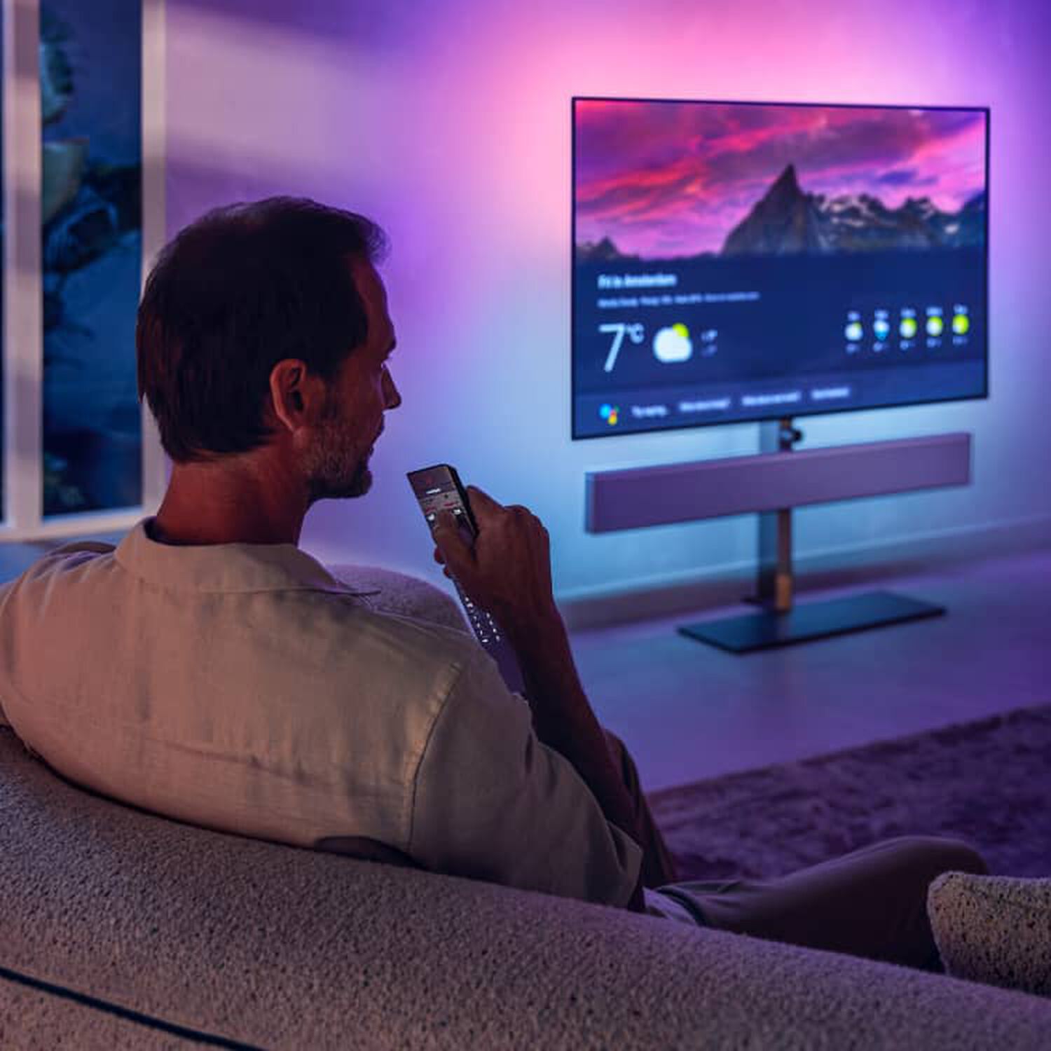 Man sitting on couch facing right, TV displaying weather with pink and blue illumination behind and silver soundbar below