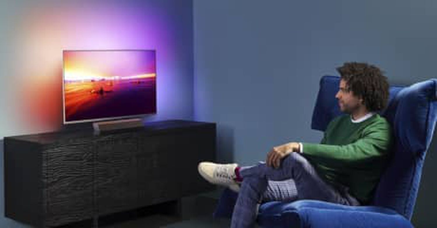 Philips OLED+9235 in living room with blue, orange, purple and white lights behind TV and man sitting in blue chair facing left looking at TV