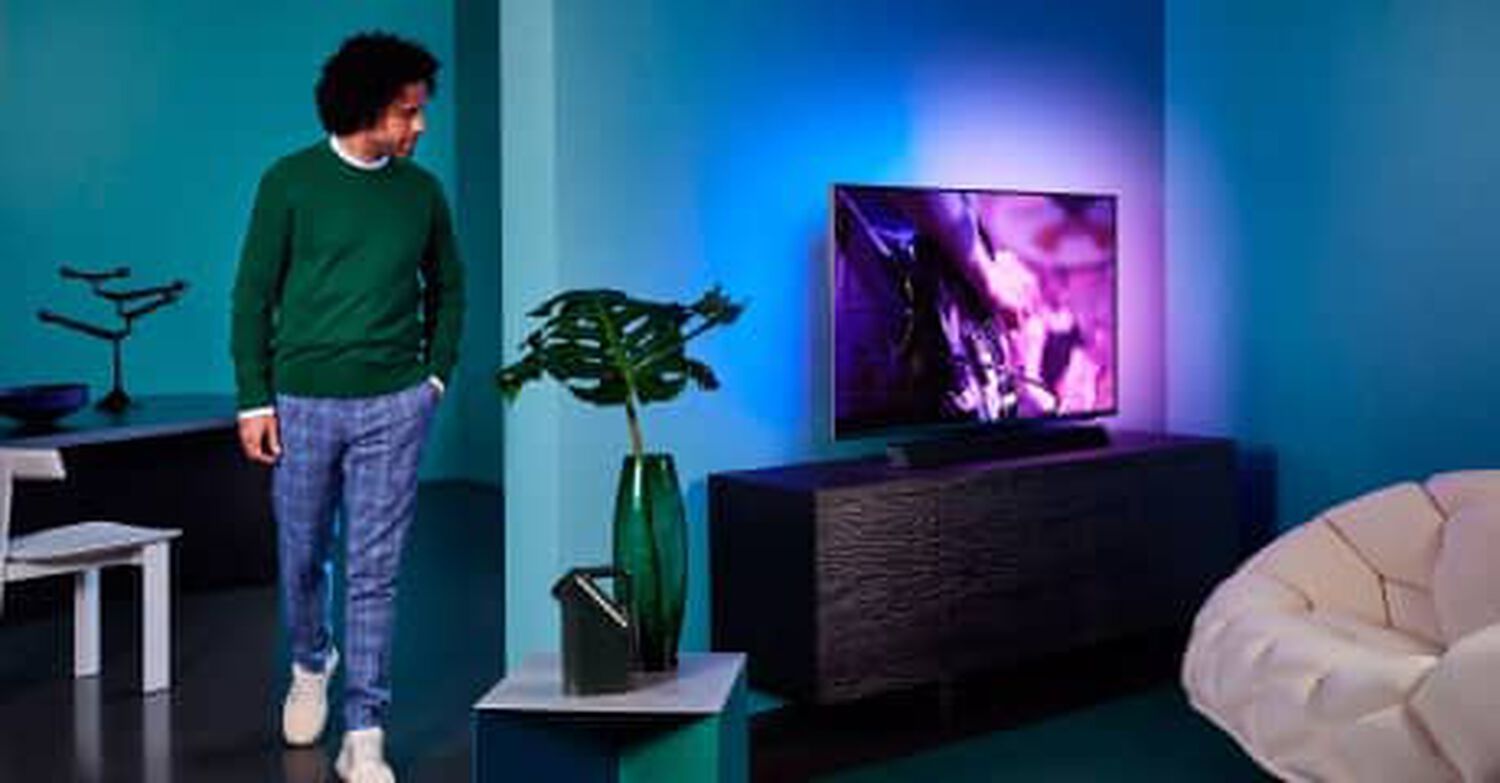 Philips OLED+9435 in living room with pink and blue lights behind TV and man facing right looking at TV in green sweater